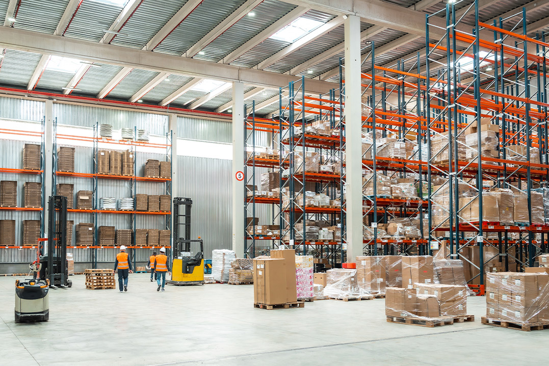 Inside a warehouse, numerous pallets are stacked, and forklifts are maneuvering, showcasing a busy logistics environment.