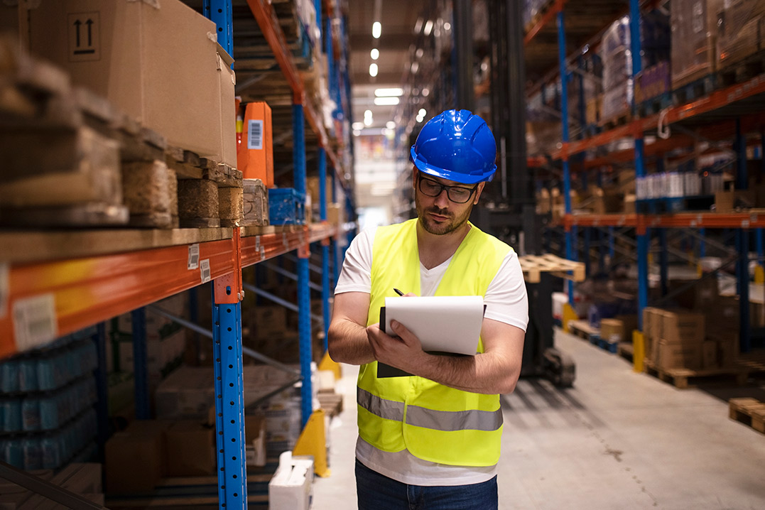 In a warehouse, a man in a safety vest and hard hat stands, ensuring safety and monitoring the environment around him.