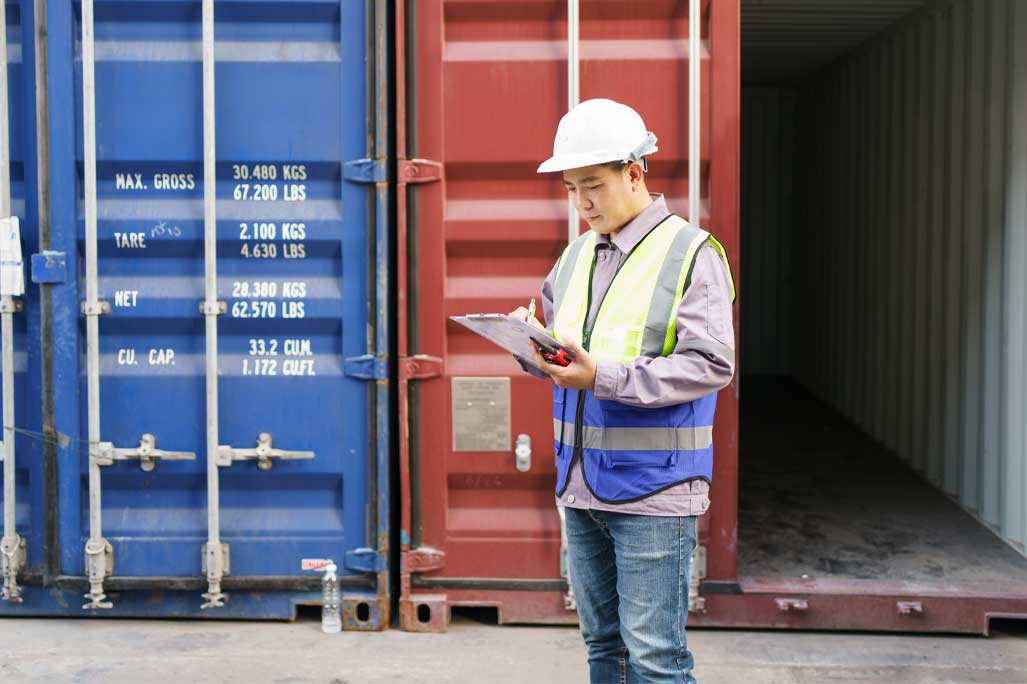 Asian male shipyard engineer is inspecting import or export cargo containers at the container yard. A customs clearance officer inspects against the import - export cargo.
