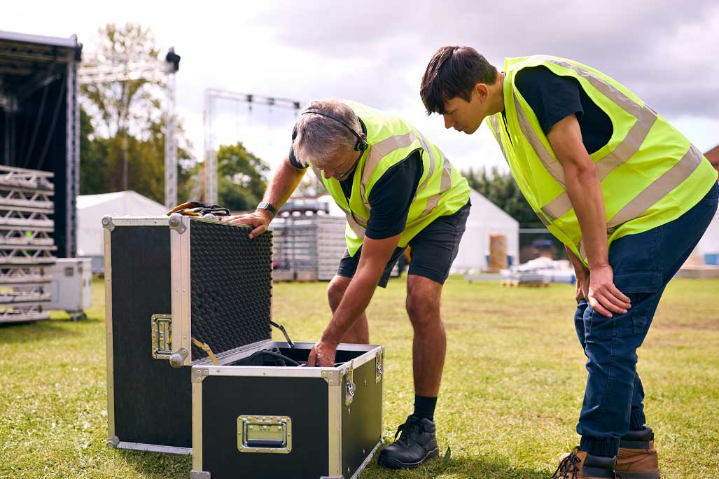 Male Production Team Unpacking Flight Case And Setting Up Outdoor Stage For Music Festival